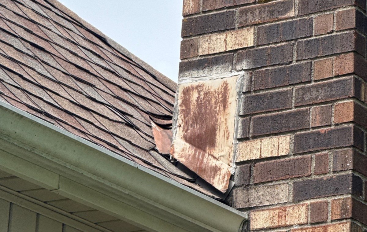The chimney flashing at inspection: rusted metal, a caulked edge, and a lifted shingle beside it.